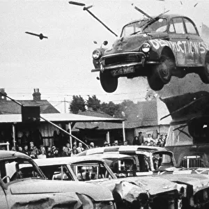 A stunt car flies through the air over a row of other cars, watched by spectators. Date: 1960s