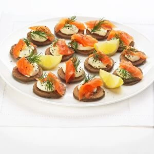 Blinis topped with smoked salmon, creme fraiche, dill and black pepper and garnished with lemon wedges, on a plate, high angle view